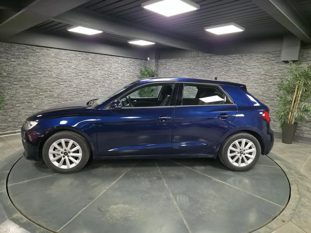 Audi A1 Sportback bleu métallisé vue de profil côté gauche dans un showroom avec sol gris et murs en pierre.