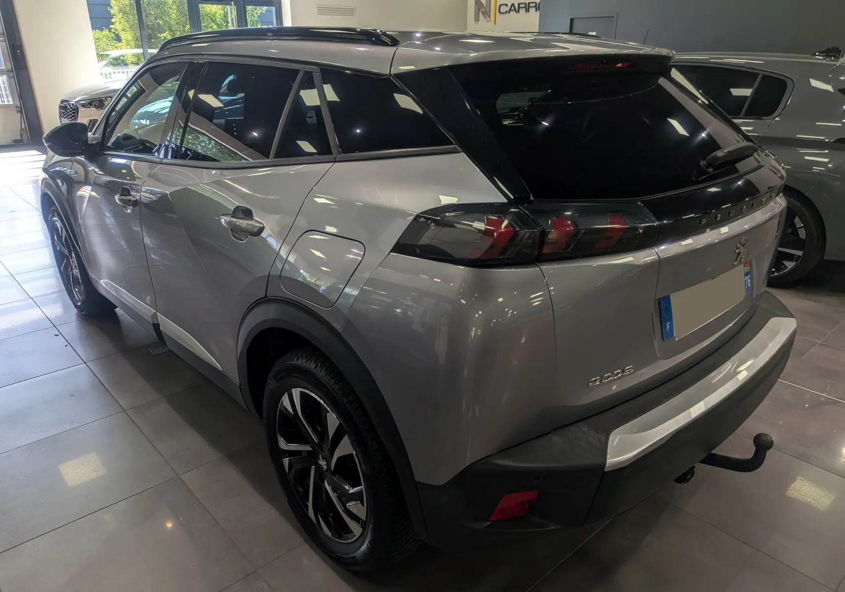 Peugeot 2008 Gris Artense vue 3/4 arrière droit, toit noir, feux arrière LED et attelage visible en showroom.