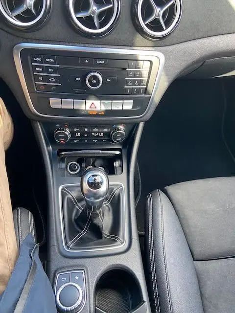 Vue intérieure centrée sur la console centrale d'une Mercedes CLA Shooting Brake 200D blanche, avec levier de vitesses manuel et aérateurs ronds chromés.