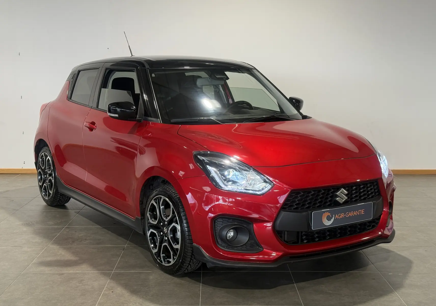 Suzuki Swift Sport rouge 3/4 avant droit avec toit noir et jantes alliage distinctives, en intérieur.