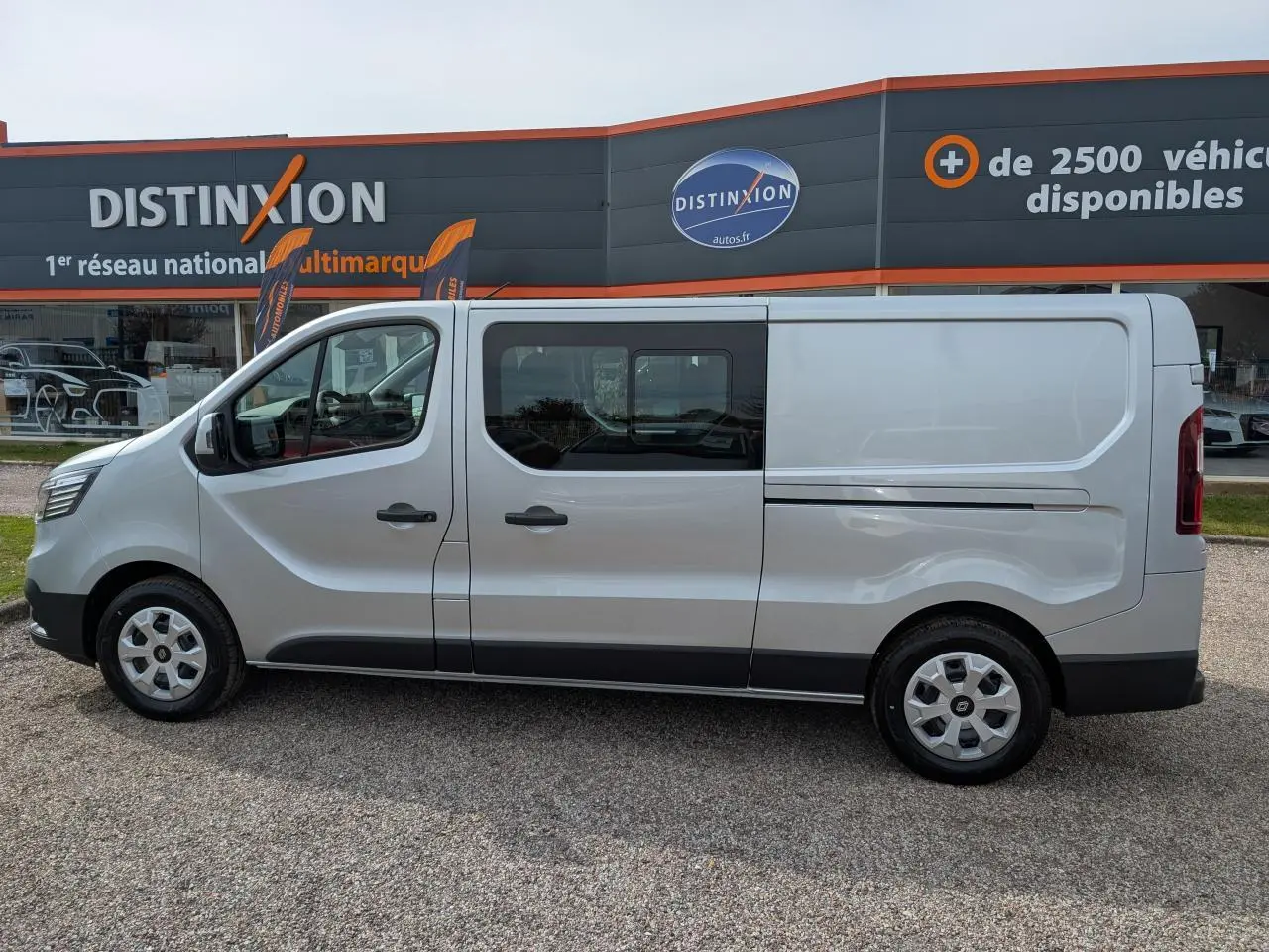 Profil côté gauche d’un Renault Trafic Fourgon gris avec cabine approfondie et enjoliveurs 16 pouces, stationné devant un concessionnaire.