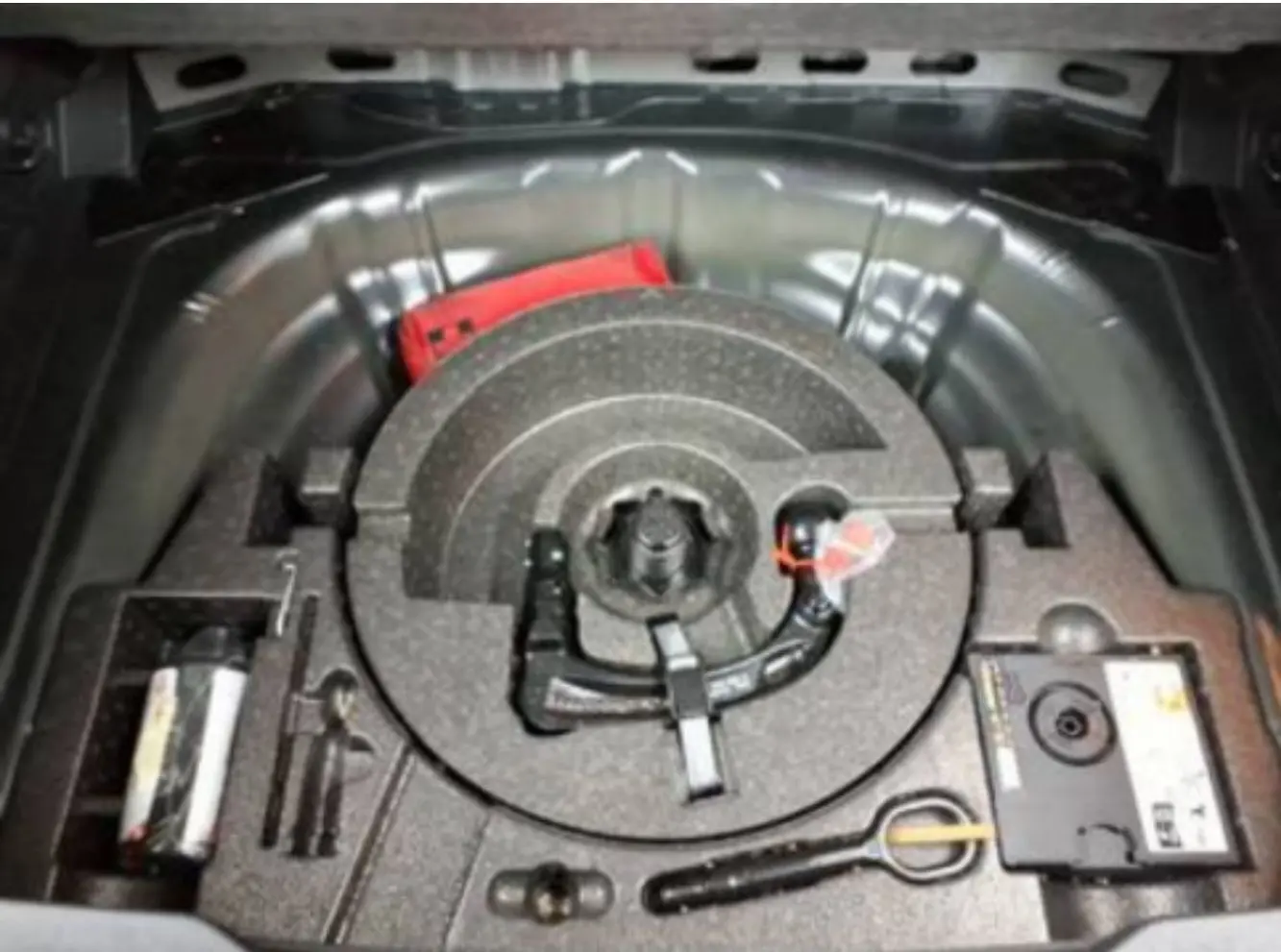 Vue plongeante sur le coffre ouvert d’un Volkswagen T-Roc noir, montrant le rangement pour roue de secours et outils.