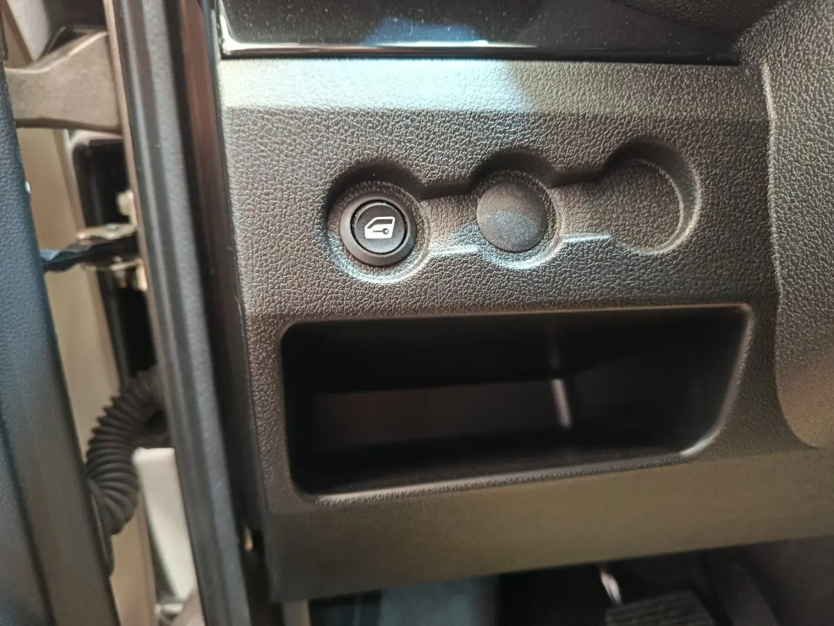 Détail intérieur montrant le bouton d'ouverture du coffre sur la partie inférieure gauche du tableau de bord d'une Aixam Ambition blanche.