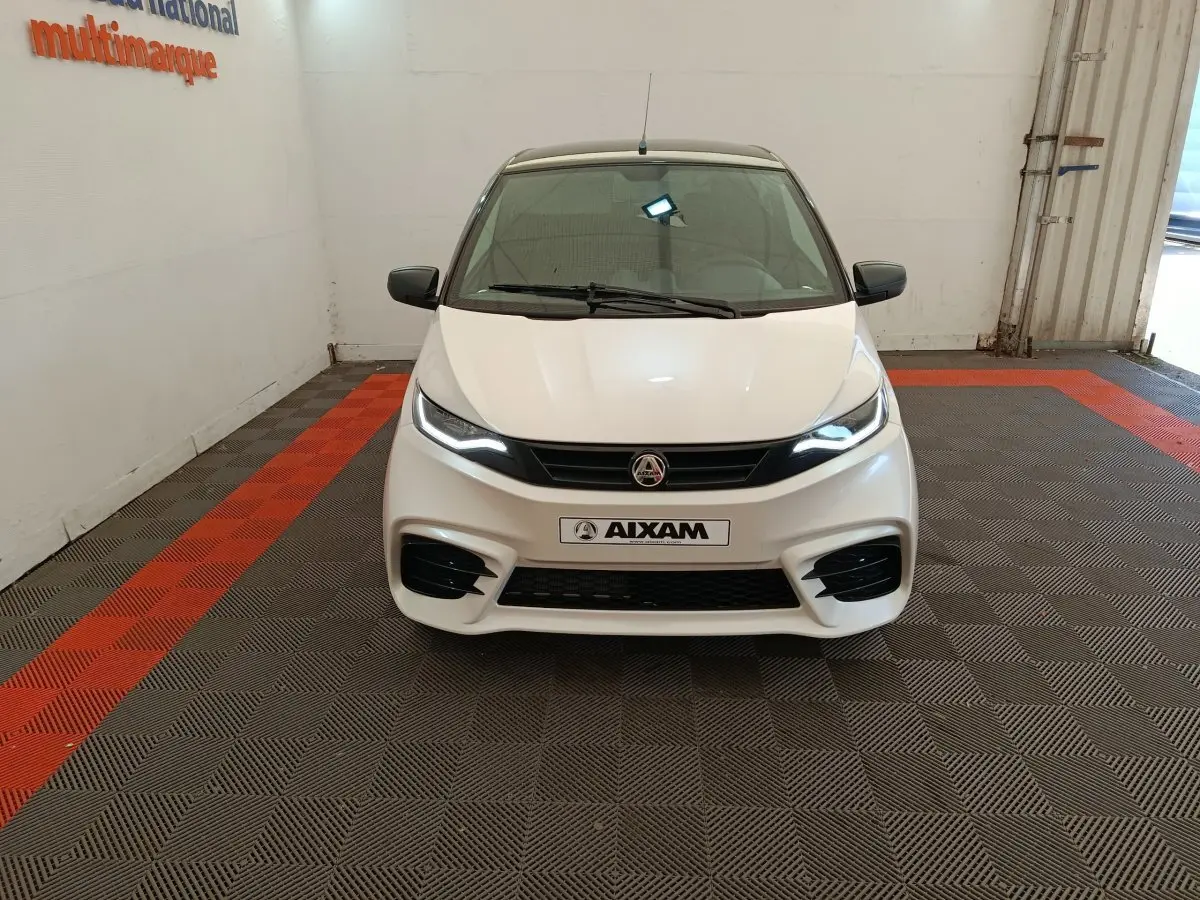 Vue avant d'une Aixam Ambition 0.5 City Coupe Sport blanche avec phares allumés et rétroviseurs noirs en intérieur showroom.
