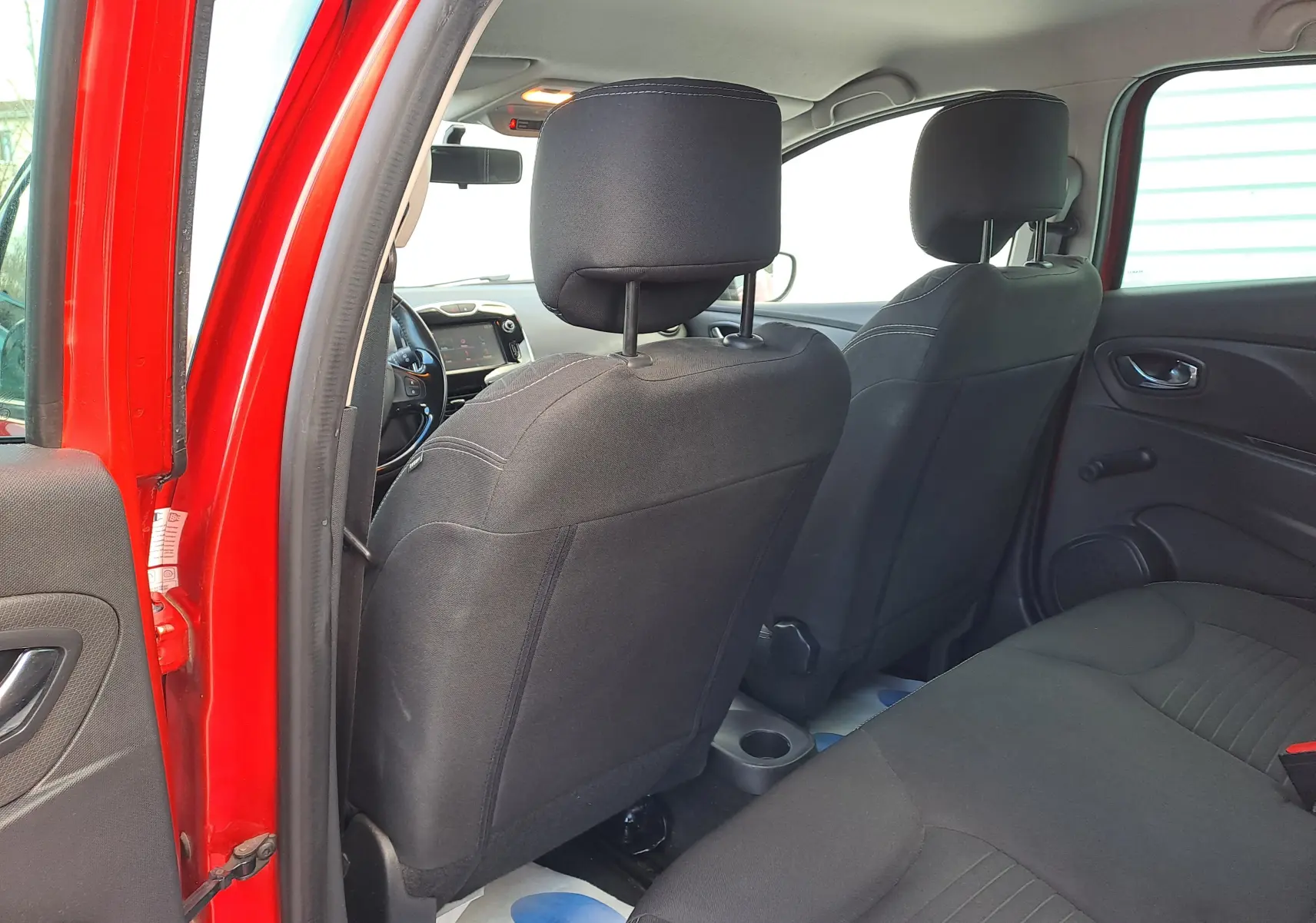 Vue intérieure arrière côté gauche de la Renault Clio IV rouge, montrant les sièges noirs et le tableau de bord à l'avant.