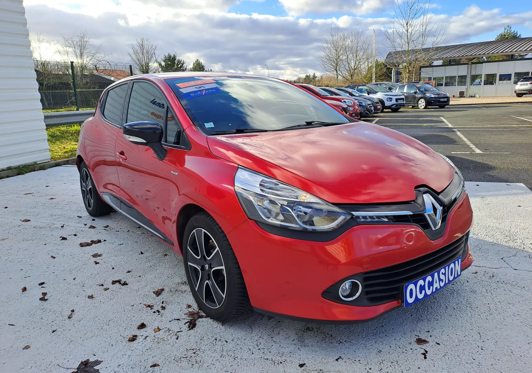Renault Clio IV rouge vue 3/4 avant droit, avec jantes noires et phares LED, stationnée sur parking extérieur.