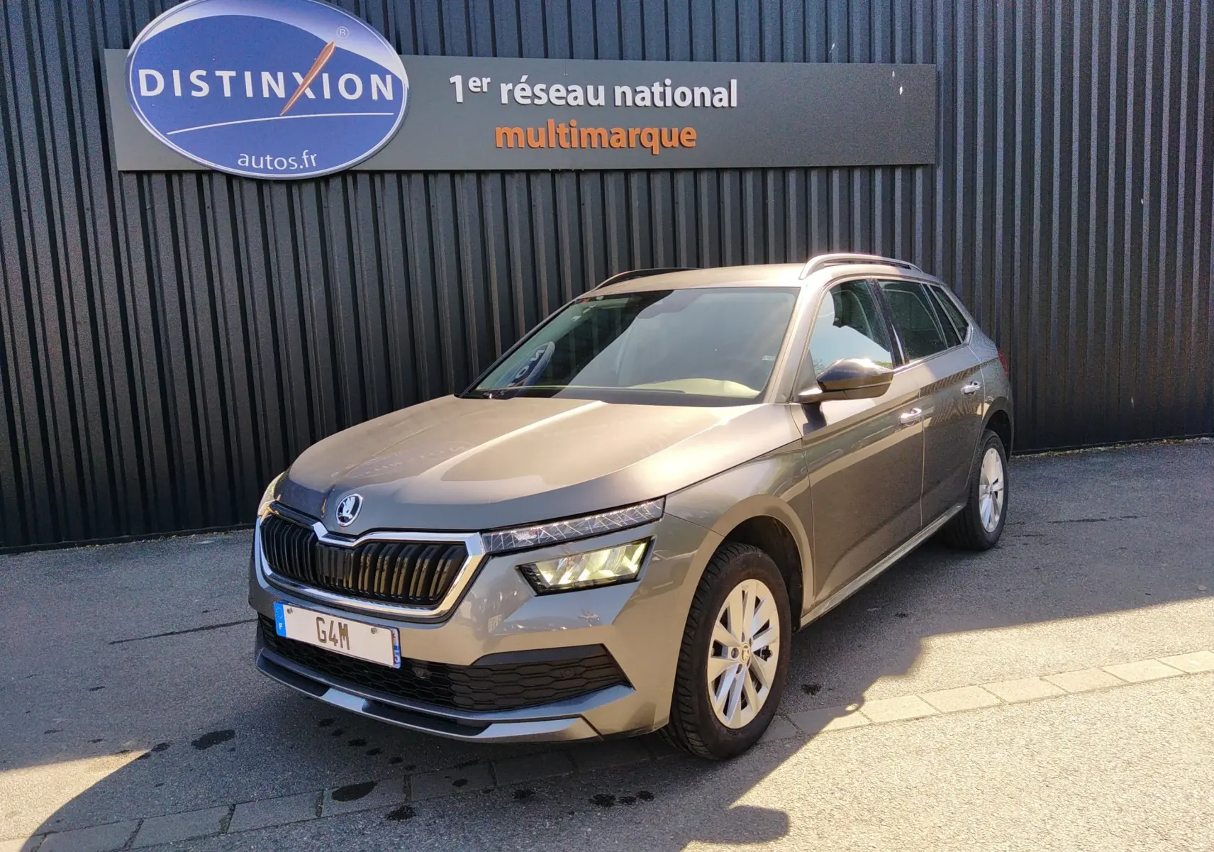 Skoda Kamiq gris 2023 en 3/4 avant droit, avec calandre noire et phares allumés, stationné devant un mur noir.
