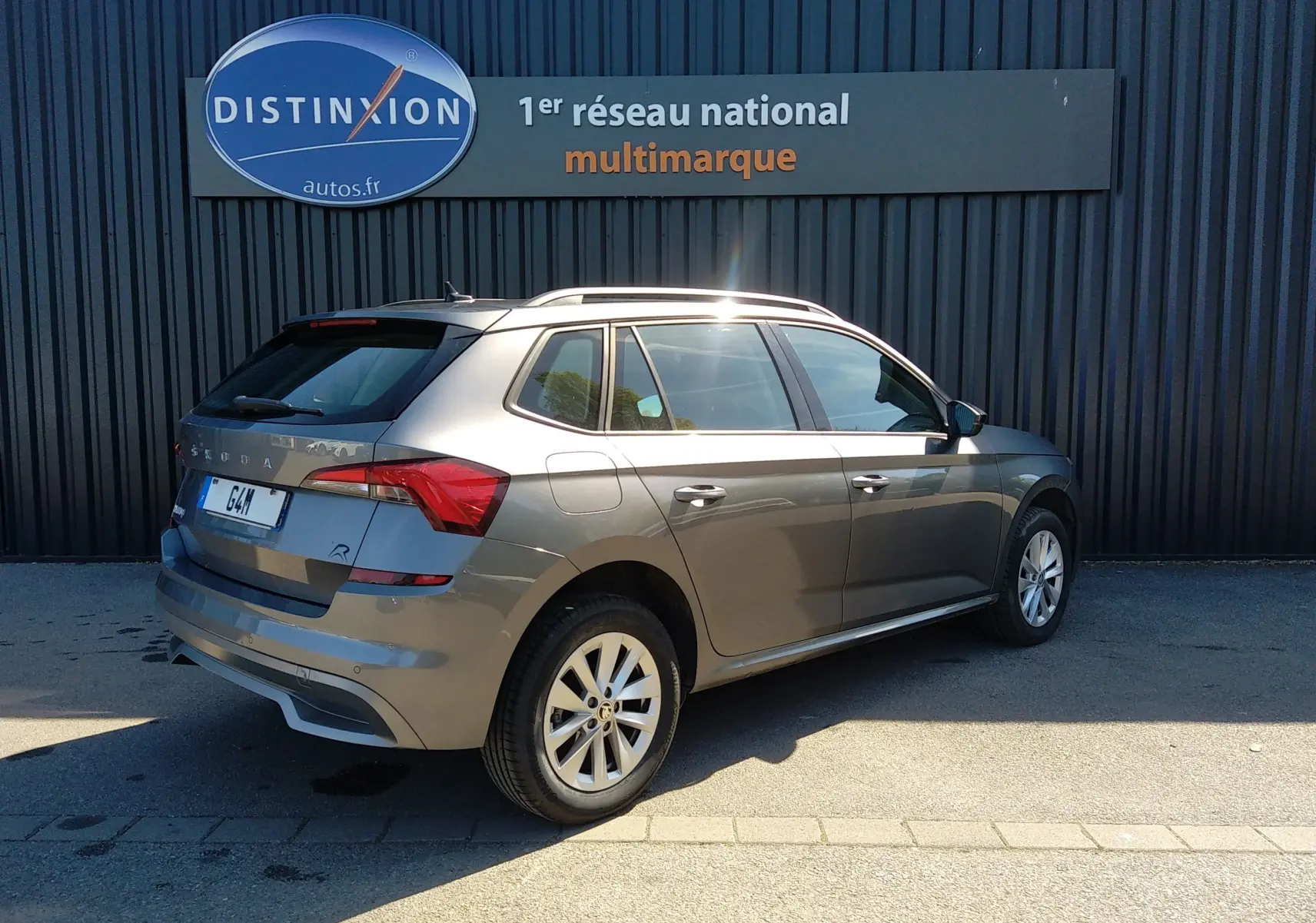 Vue 3/4 arrière droite d'un Skoda Kamiq gris 2023 avec jantes alliage et toit ouvrant, stationné devant un mur noir.