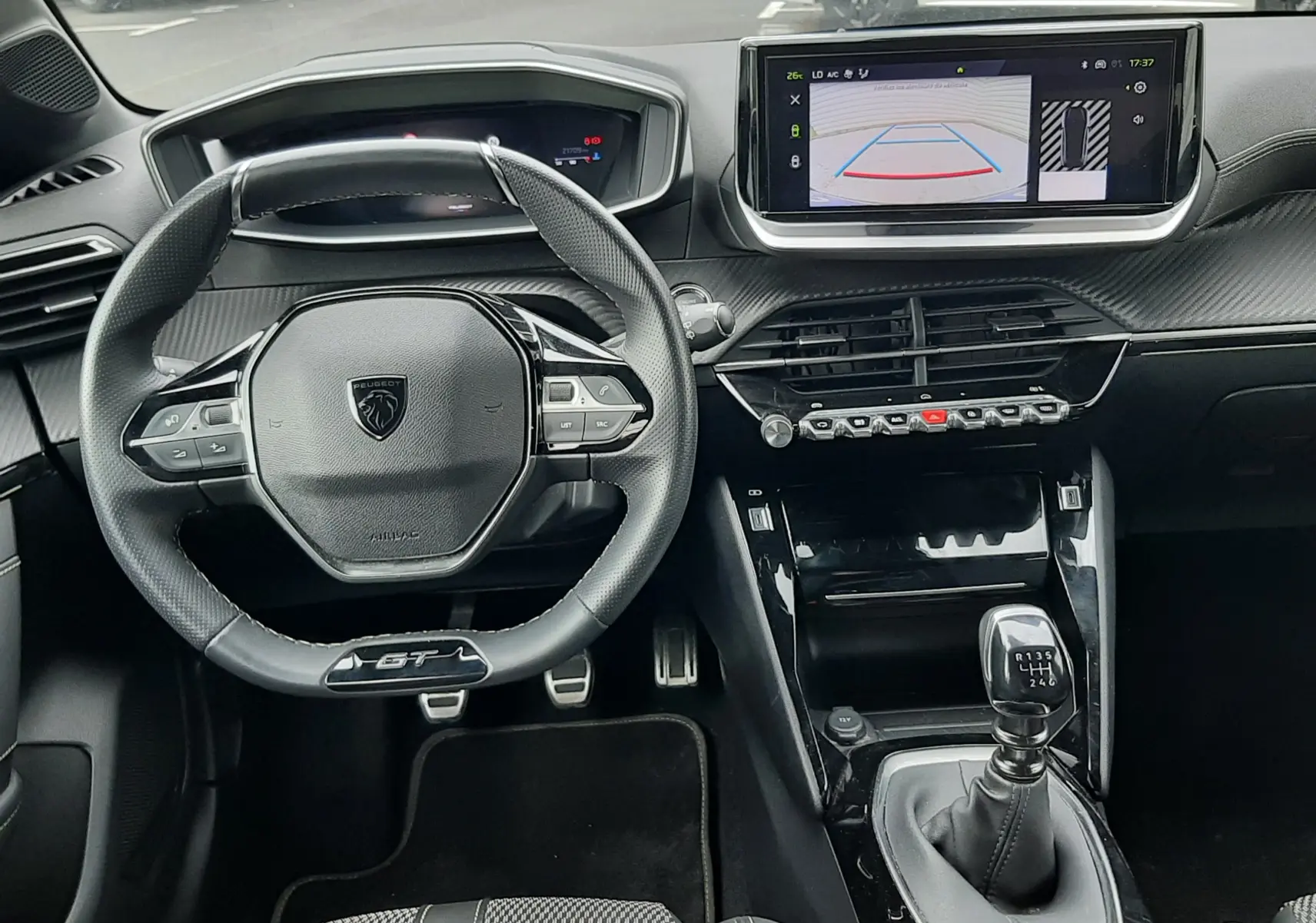 Vue intérieure avant de la Peugeot 208 GT 1.2 essence 2025, volant GT, tableau de bord numérique et écran tactile avec caméra de recul.