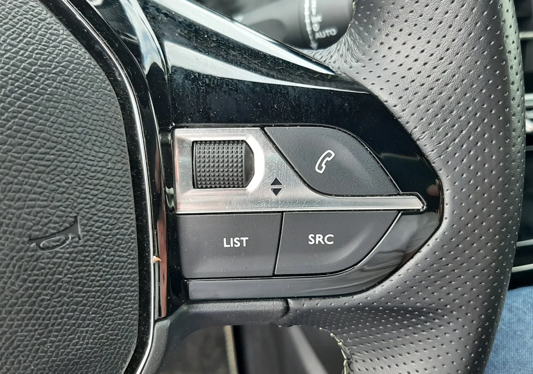 Gros plan sur les commandes gauche du volant perforé noir de la Peugeot 208 gris Artense, boutons LIST et SRC visibles.