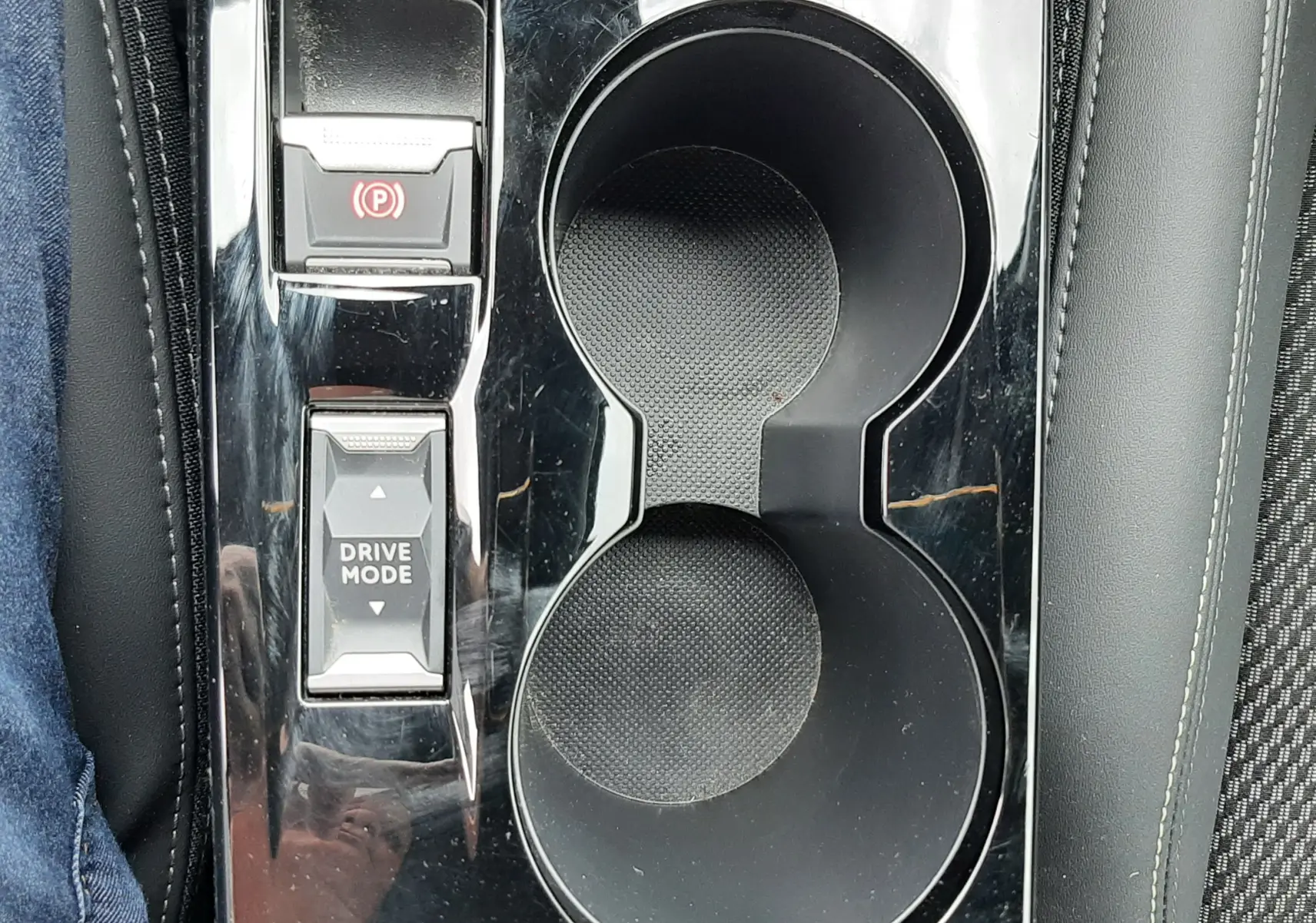 Gros plan sur la console centrale noire brillante de la Peugeot 208 1.2 100CH, avec porte-gobelets et boutons de frein à main et mode de conduite.