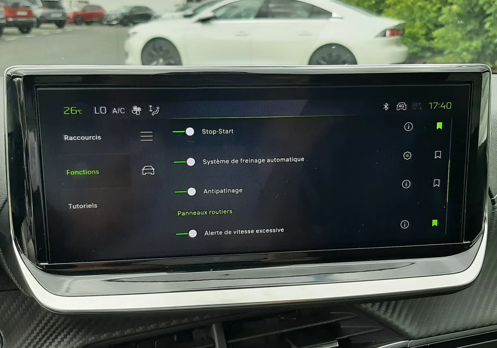 Vue intérieure centrée sur l'écran tactile du tableau de bord, avec en arrière-plan une Peugeot 208 gris Artense vue de profil côté gauche.