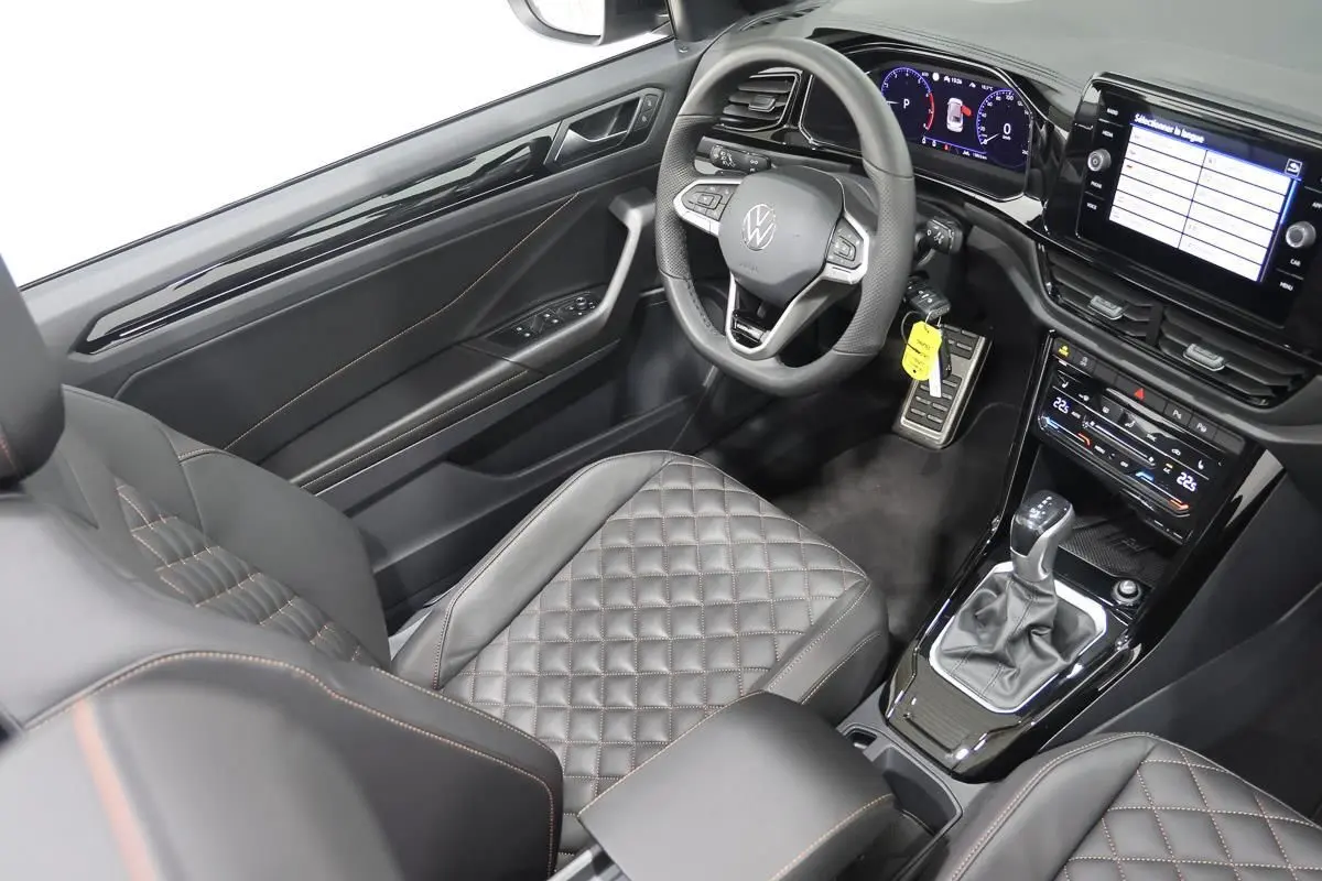 Vue intérieure côté conducteur du Volkswagen T-Roc Cabriolet 2025, sièges cuir noir matelassé avec surpiqûres orange.