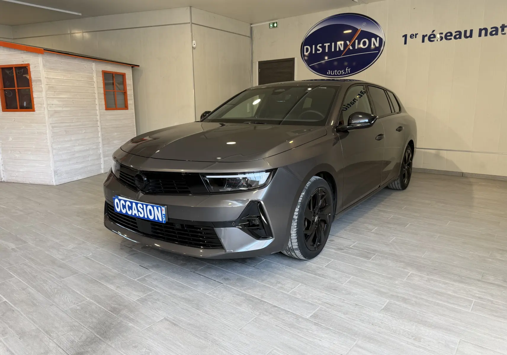 Opel Astra Sports Tourer gris vue 3/4 avant droit en intérieur avec jantes noires et plaque occasion.