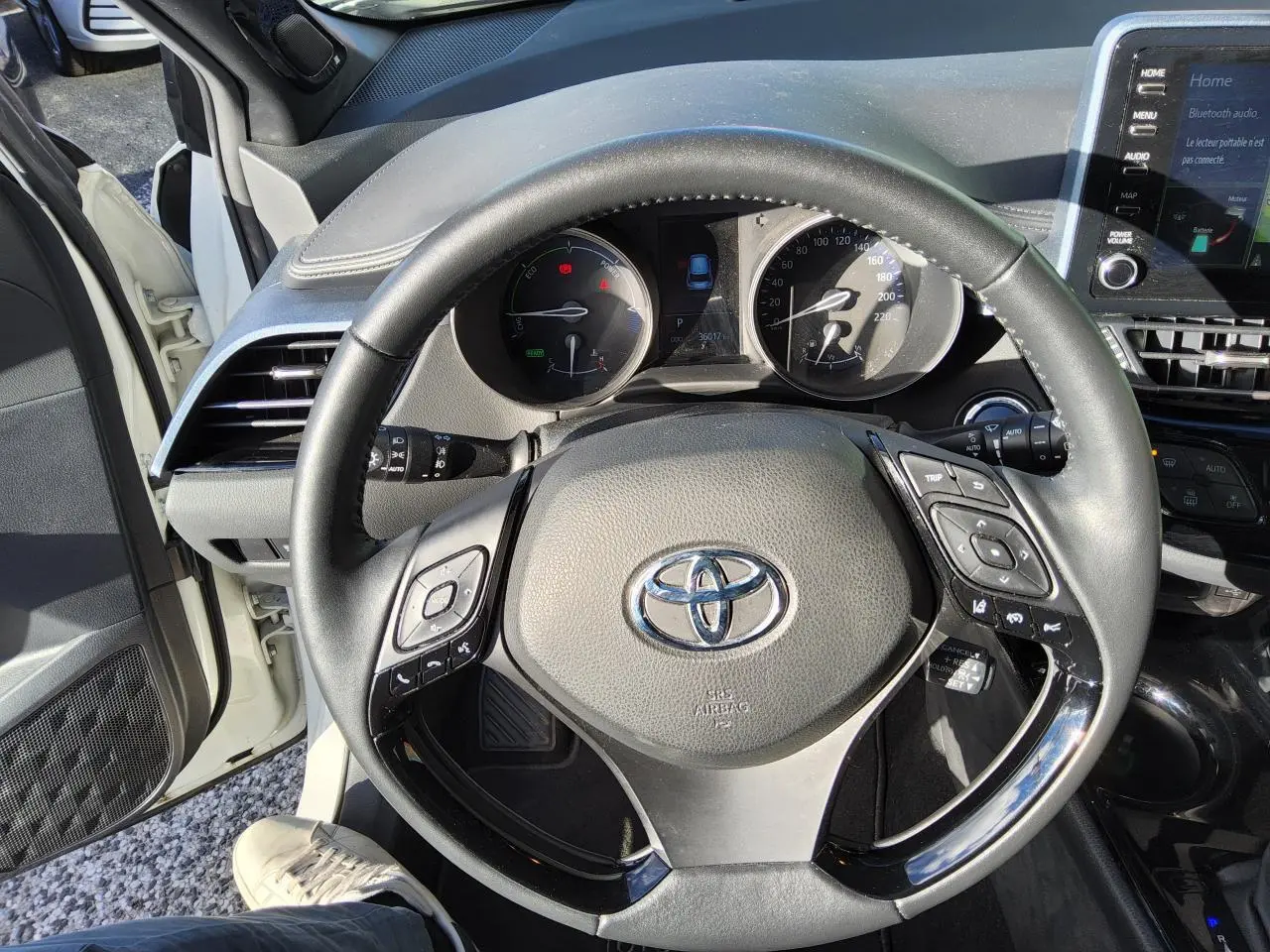 Vue intérieure centrée sur le volant noir du Toyota C-HR blanc nacré, avec tableau de bord et écran tactile visibles.