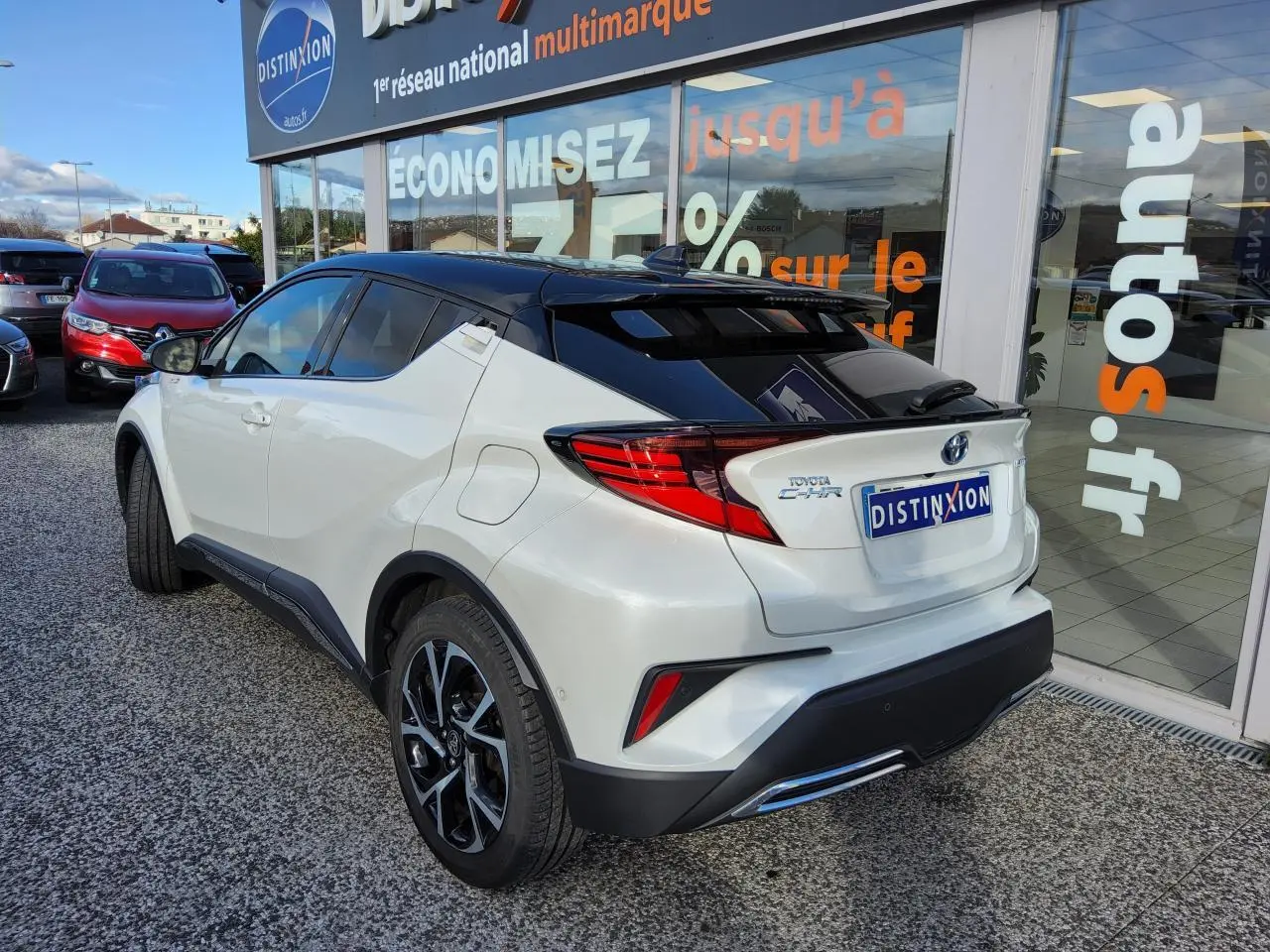 Toyota C-HR blanc nacré en 3/4 arrière droit, toit noir et jantes alliage 18 pouces visibles devant un showroom.