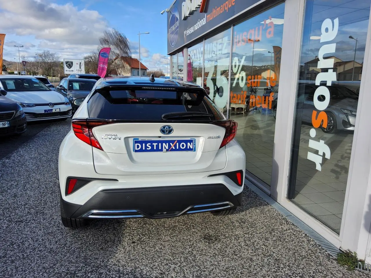 Vue arrière d'un Toyota C-HR blanc nacré avec ornements chromés, stationné devant une vitrine de concession multimarque.