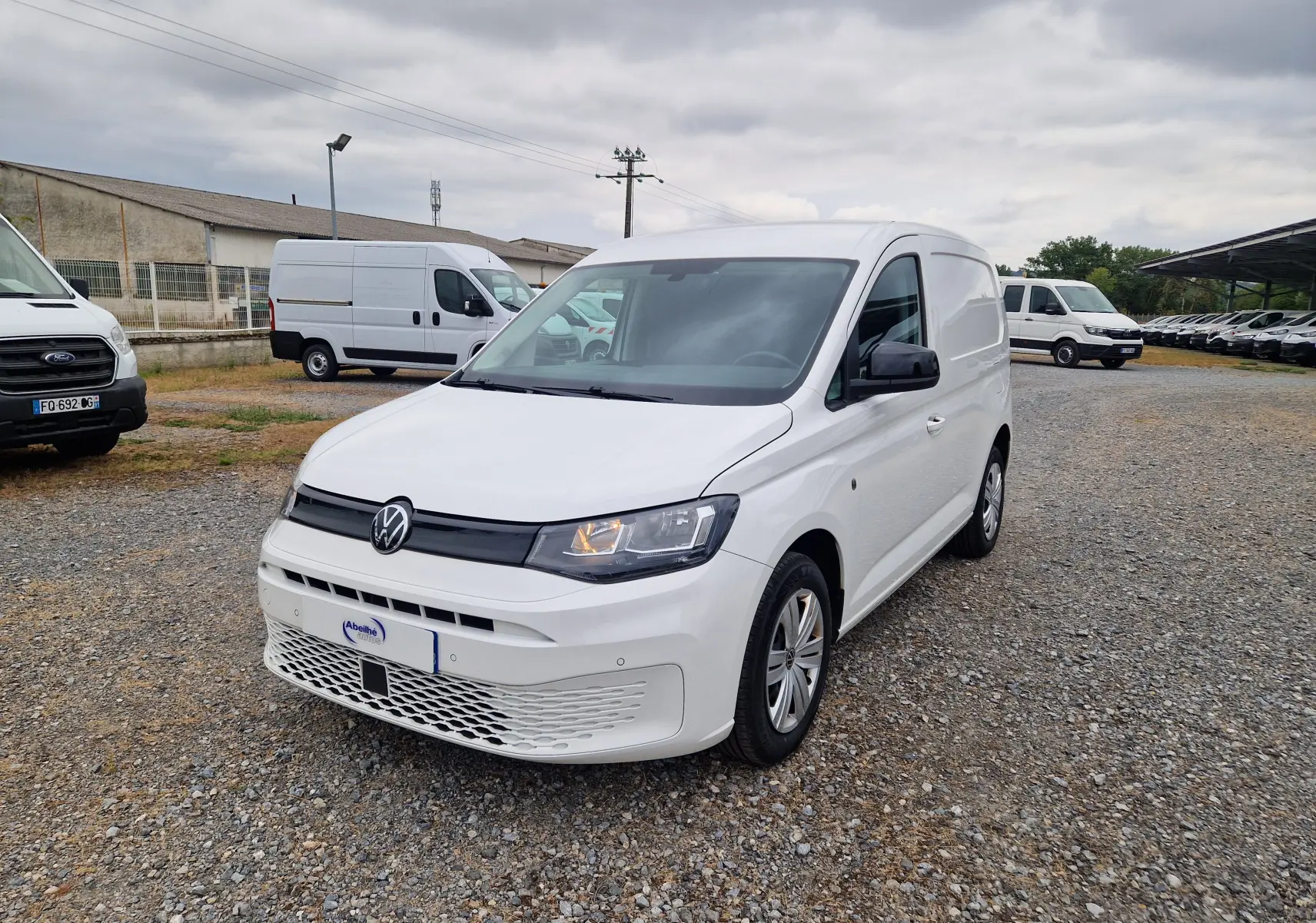 Volkswagen Caddy Cargo blanc vu en 3/4 avant droit sur un sol gravillonné avec plusieurs utilitaires en arrière-plan.
