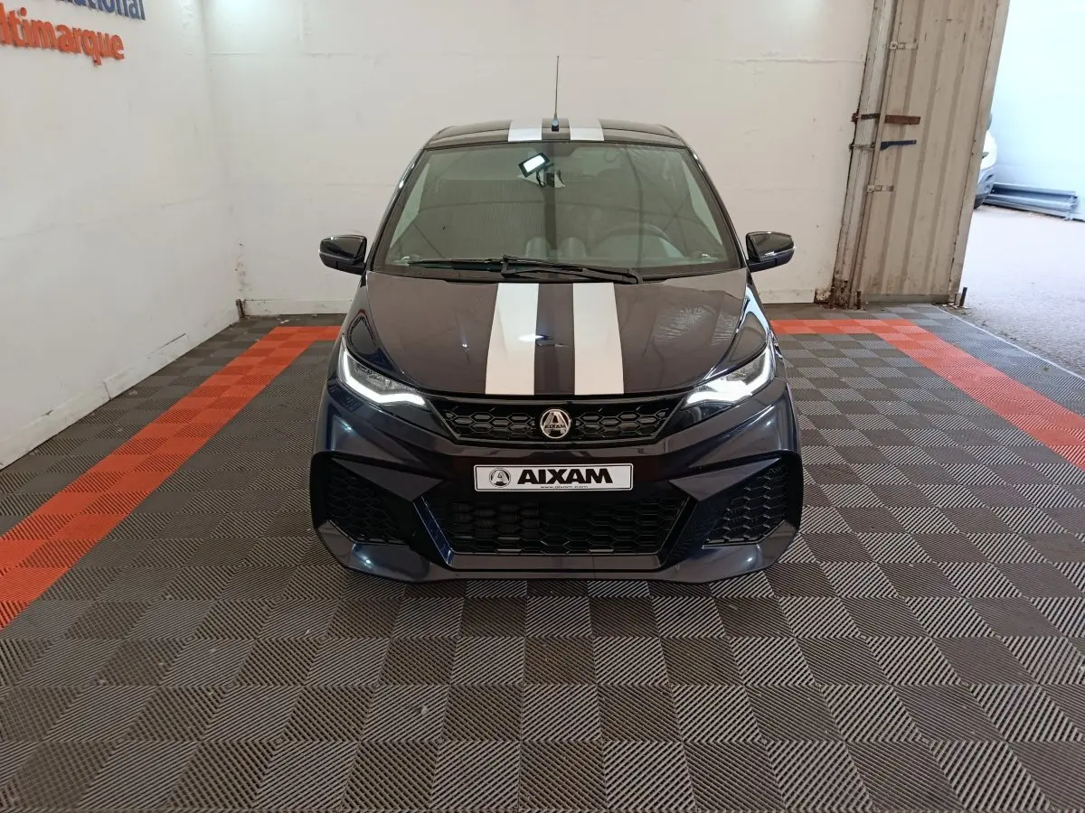 Vue avant d'une Aixam Ambition 0.5 City Coupe GTI noire avec bandes blanches sur le capot, phares allumés, en intérieur.