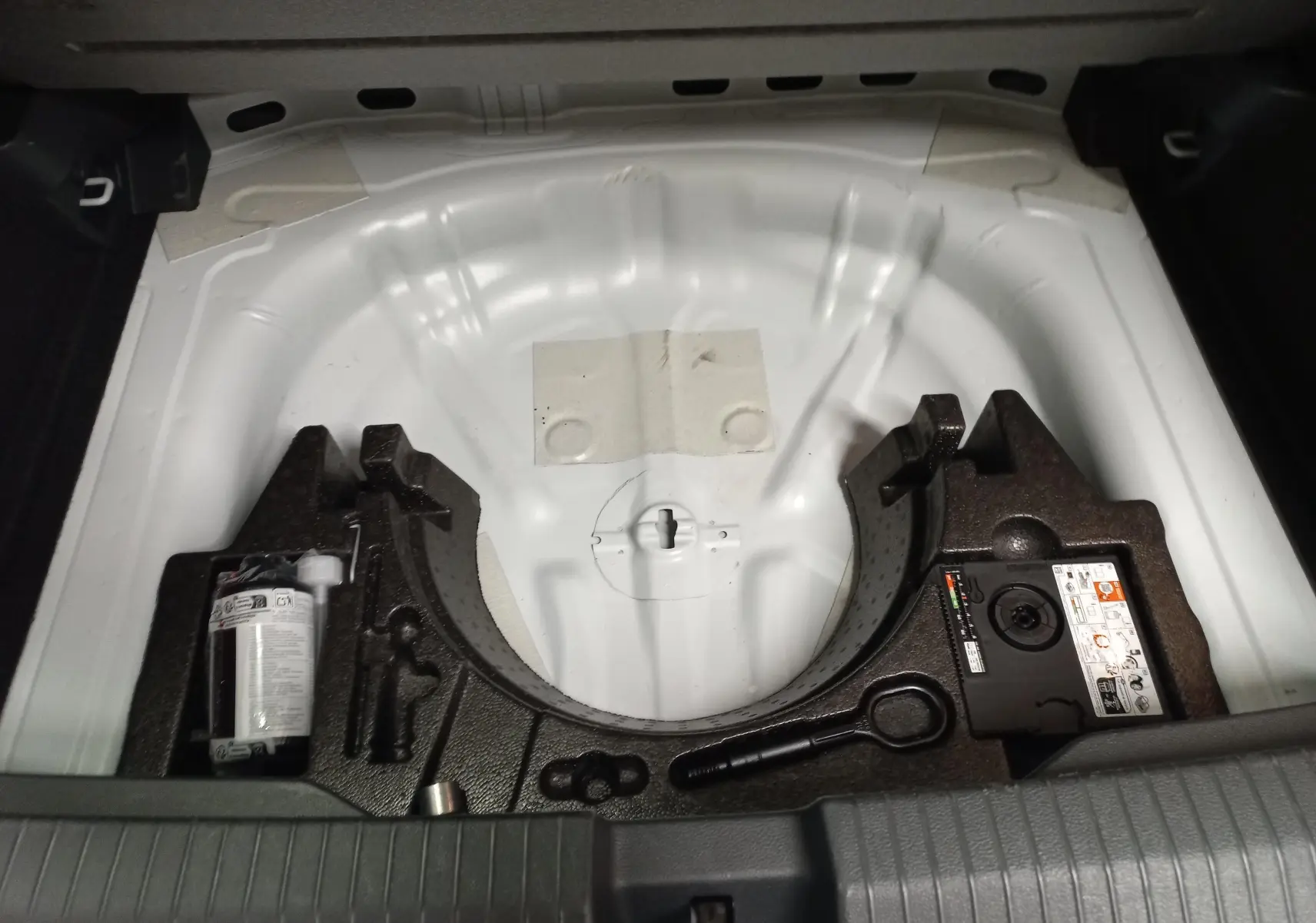 Vue plongeante sur le coffre ouvert d'un Volkswagen T-Roc blanc pur, montrant le rangement pour roue de secours et outils.