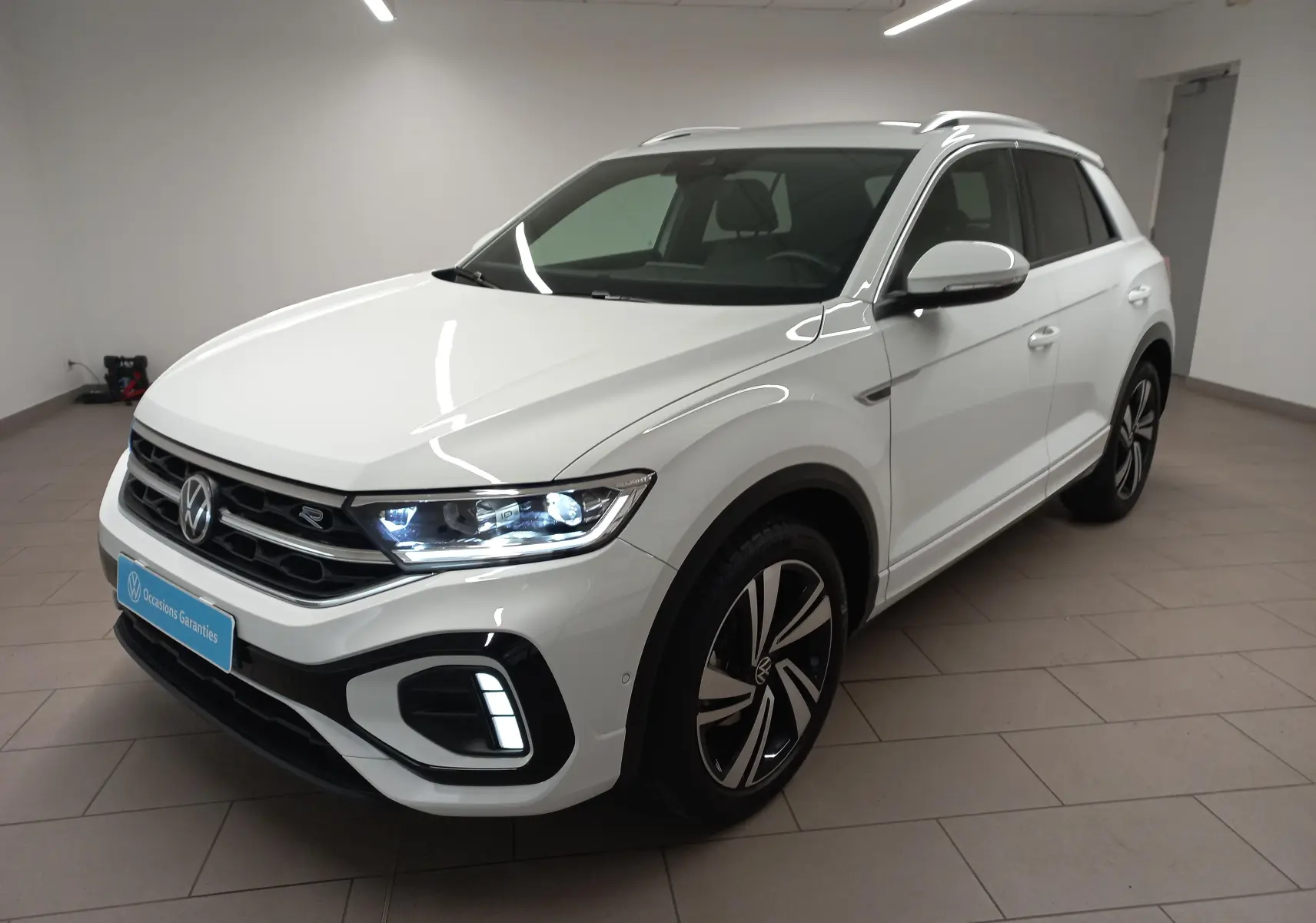 Vue 3/4 avant droit du Volkswagen T-Roc blanc pur 2025 R-Line avec feux LED allumés en intérieur.