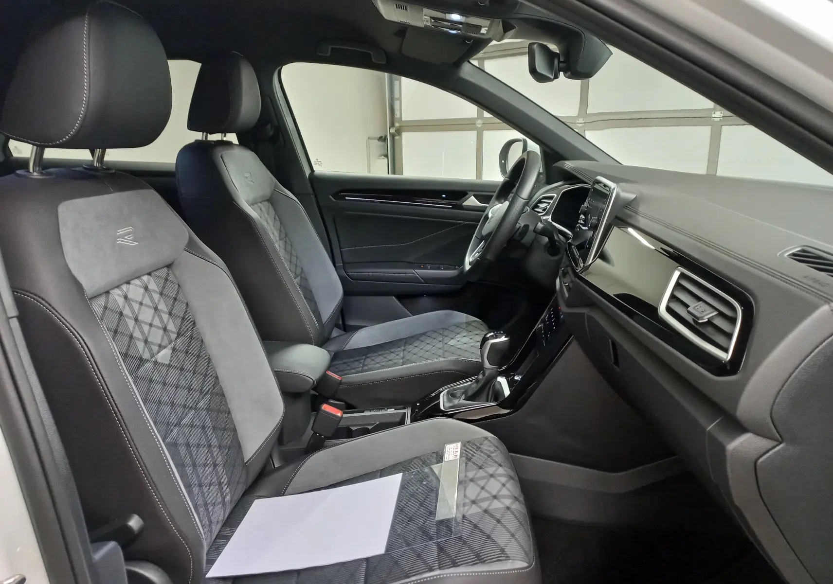 Vue intérieure côté gauche du Volkswagen T-Roc 1.5 TSI 150 DSG7 R-Line Edition, sièges noirs avec surpiqûres et tableau de bord moderne.