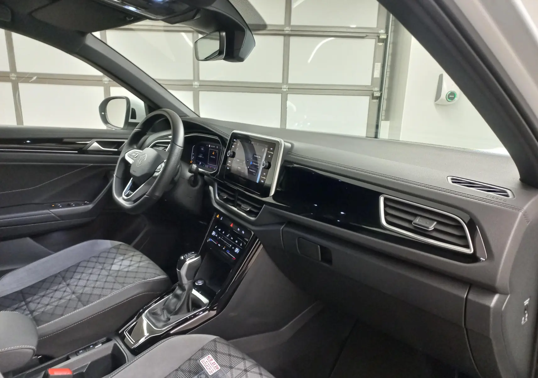 Vue intérieure côté conducteur du Volkswagen T-Roc 1.5 TSI blanc, avec tableau de bord moderne et sièges tissu à motifs.