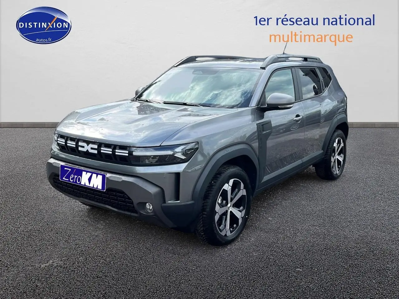 Vue 3/4 avant du Dacia Duster gris schiste 2025 avec jantes alu et barres de toit visibles sur sol bitumé.