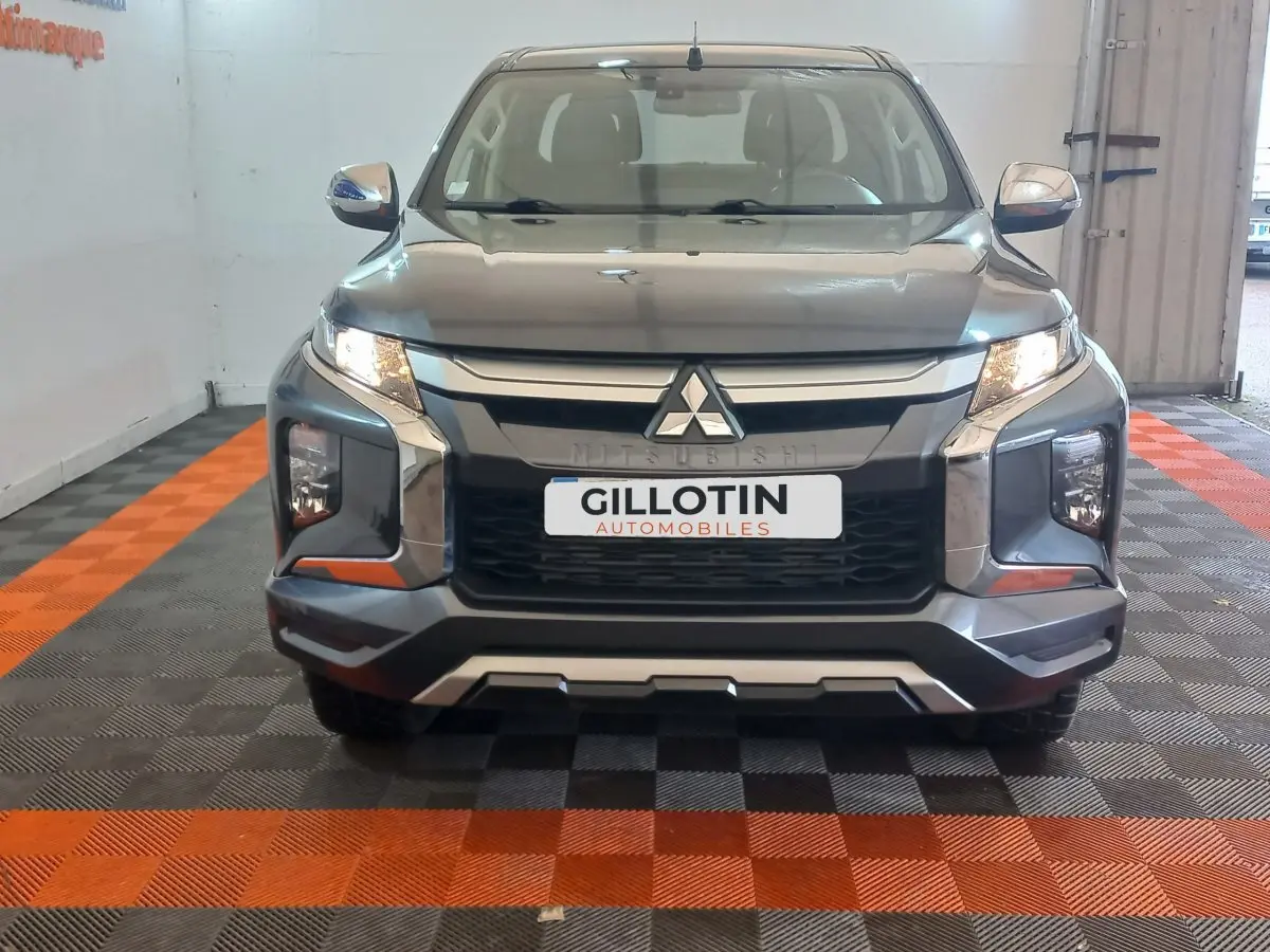Vue frontale d'un Mitsubishi L200 gris foncé avec calandre chromée et phares allumés en intérieur showroom.