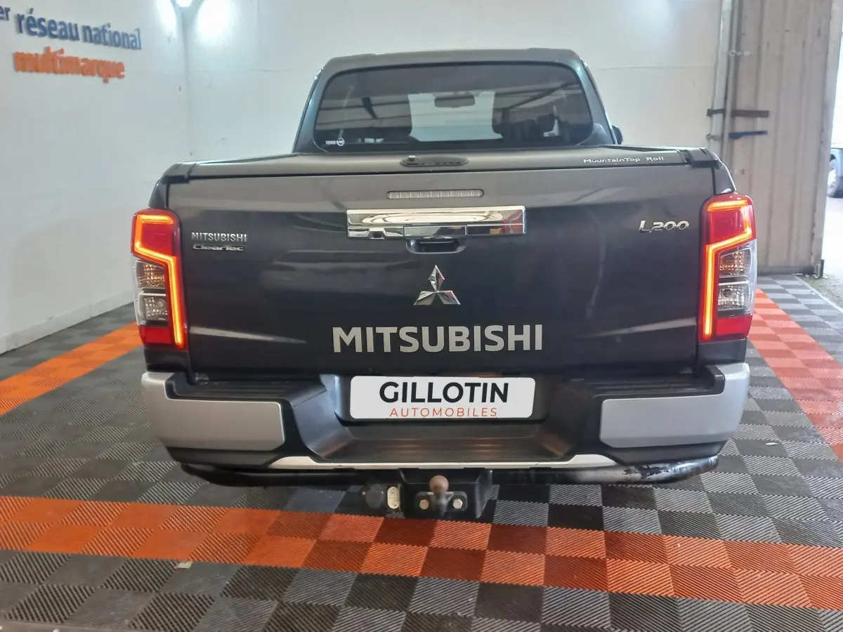 Vue arrière d’un pick-up Mitsubishi L200 gris foncé avec feux allumés et attelage visible en intérieur showroom.