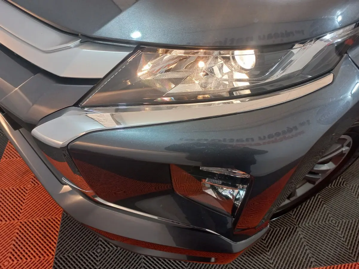 Gros plan sur l'angle avant droit du Mitsubishi L200 gris foncé, mettant en valeur le phare allumé et la calandre chromée.