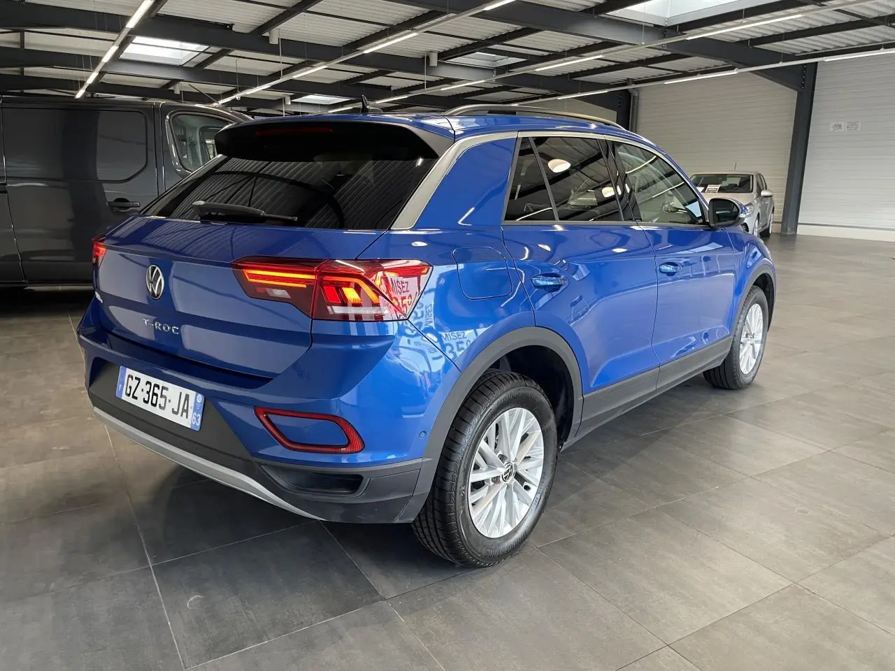 Vue 3/4 arrière droite d'un Volkswagen T-Roc bleu avec feux arrière LED allumés dans un garage.