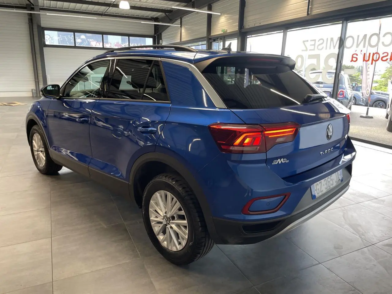 Vue 3/4 arrière droite d'un Volkswagen T-Roc bleu avec feux arrière allumés et jantes alliage en showroom.