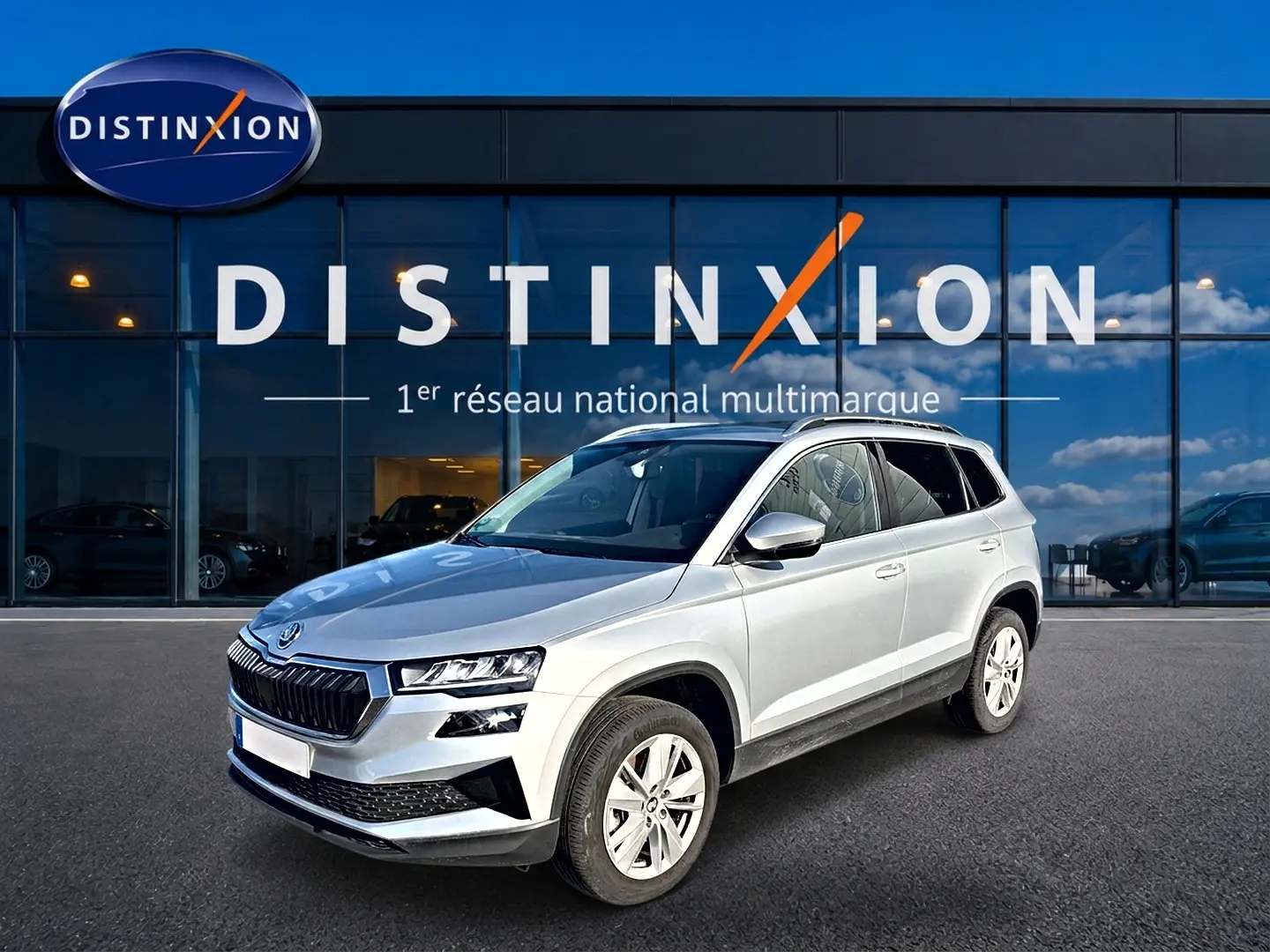 Vue 3/4 avant droite d'un Skoda Karoq gris 2025 devant une vitrine Distinxion, avec calandre noire et jantes alu.