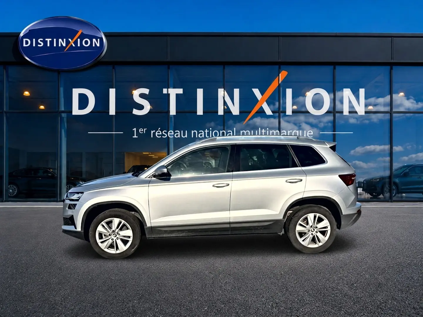 Vue de profil côté gauche d'un Skoda Karoq gris 2025 garé devant une vitrine avec logo Distinxion.