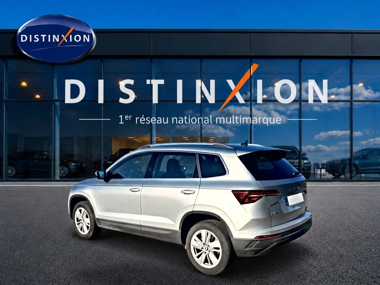 Vue 3/4 arrière droite d’un Skoda Karoq gris 2025 garé devant une vitrine avec logo Distinxion.
