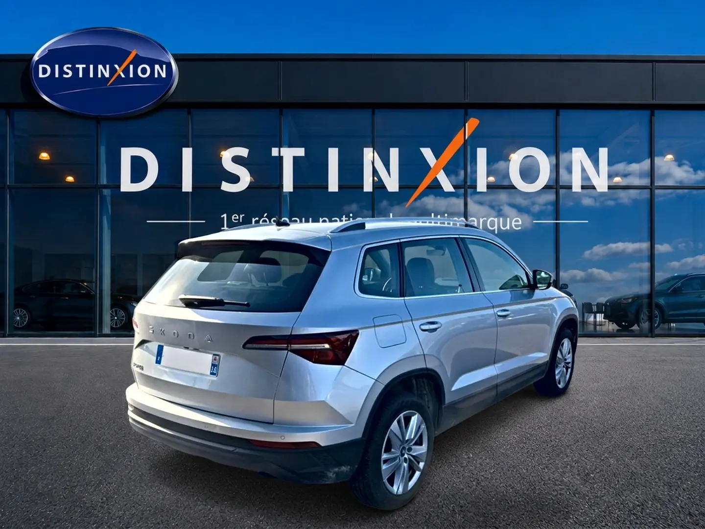 Vue 3/4 arrière droite d’un Skoda Karoq gris devant une vitrine avec logo Distinxion.