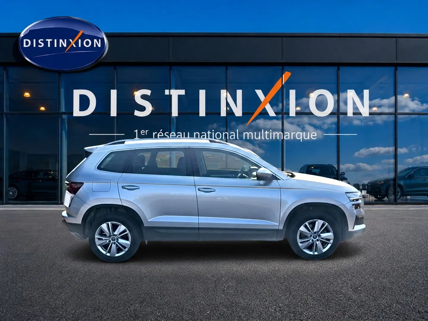 Profil droit d'un Skoda Karoq gris 2025 garé devant une vitrine avec logo Distinxion visible en arrière-plan.