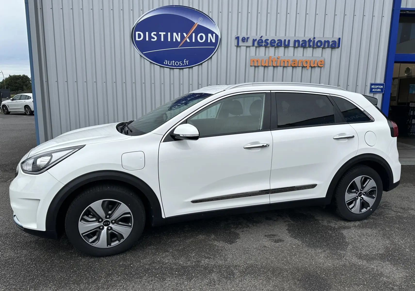 Vue de profil côté gauche d'un KIA Niro blanc céleste hybride rechargeable avec jantes spécifiques et prise de recharge visible.