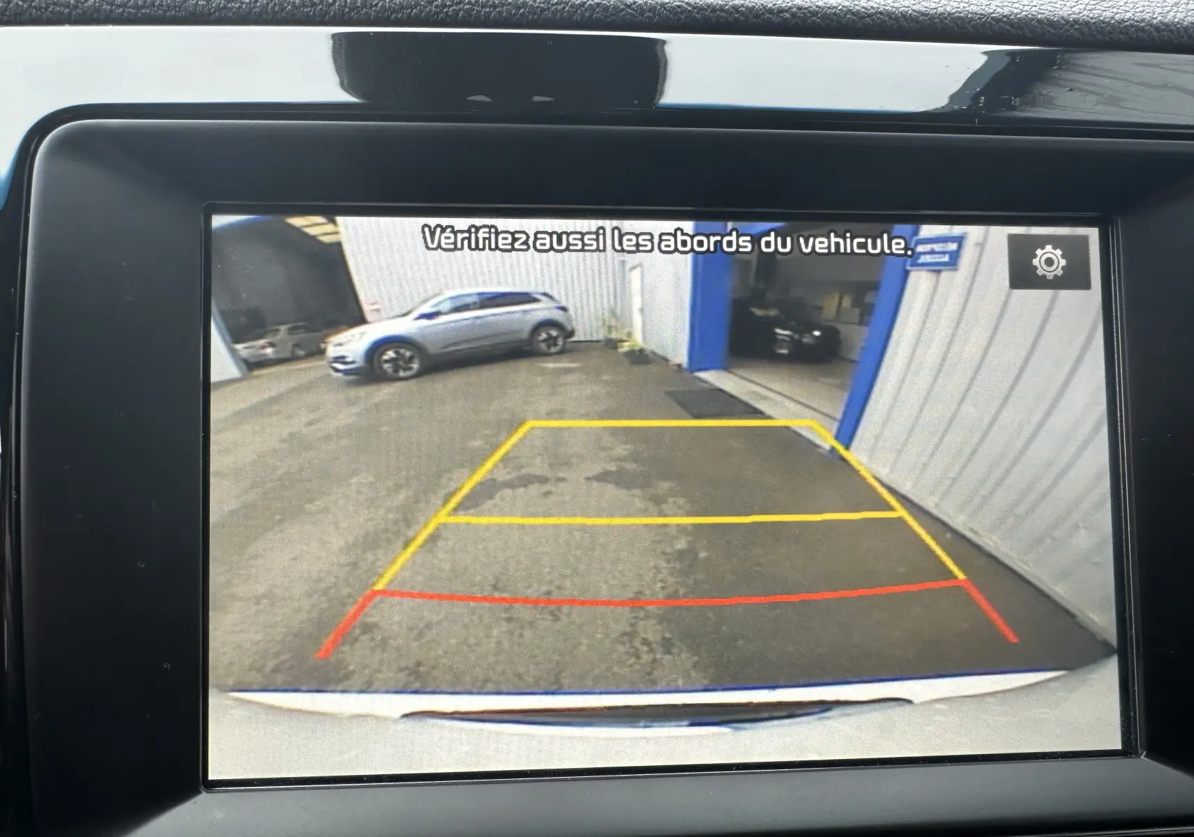 Vue caméra de recul montrant l'arrière blanc céleste du KIA NIRO 2020 et une voiture grise en stationnement à gauche.