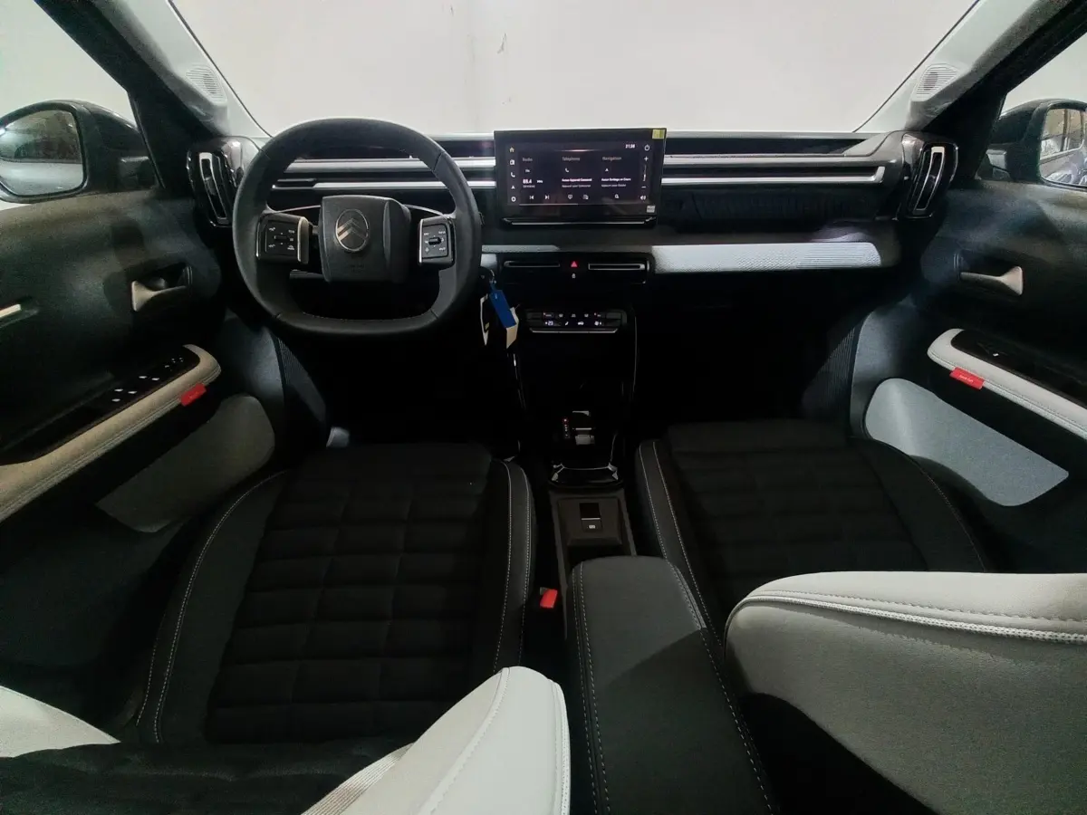 Vue intérieure avant de la Citroën C3 hybride 2025, tableau de bord noir avec écran tactile central et sièges bicolores.