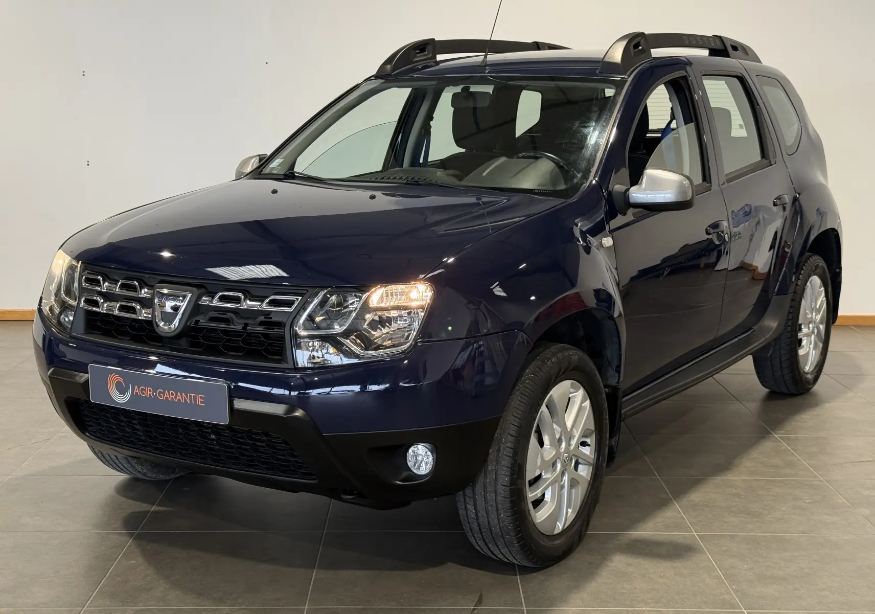 Dacia Duster bleu en 3/4 avant droit, avec calandre chromée et phares allumés dans un intérieur neutre.