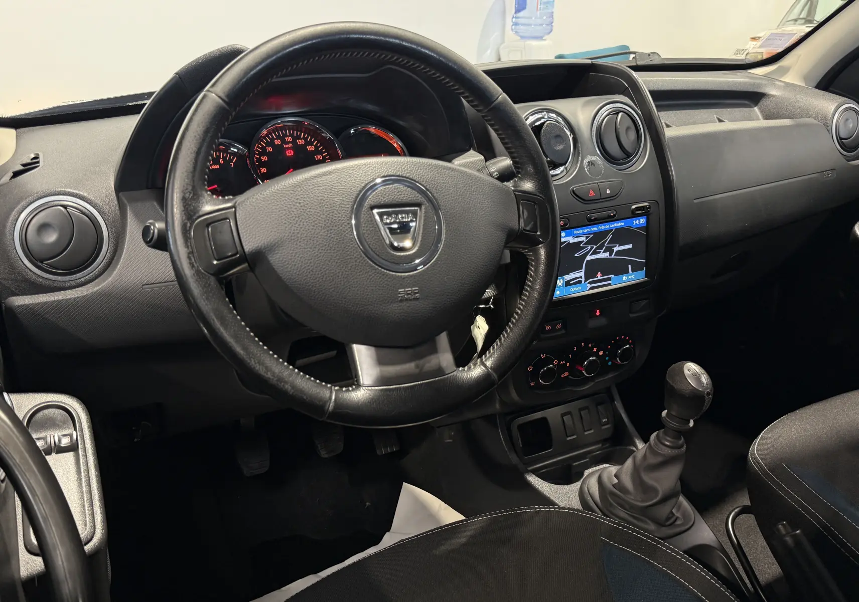 Vue intérieure du tableau de bord du Dacia Duster 2015, avec volant noir, écran tactile et levier de vitesse manuel.