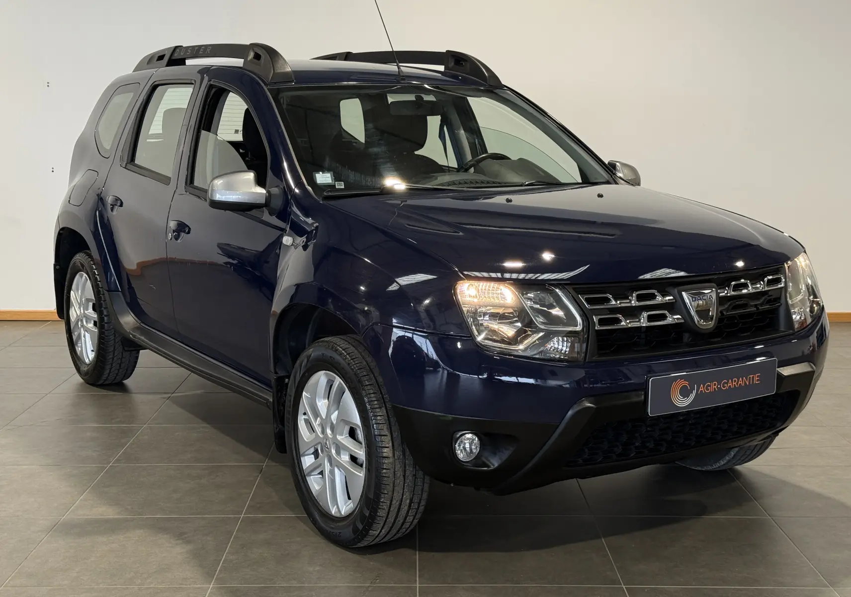 Dacia Duster bleu en 3/4 avant droit, avec calandre chromée et barres de toit noires visibles en intérieur showroom.