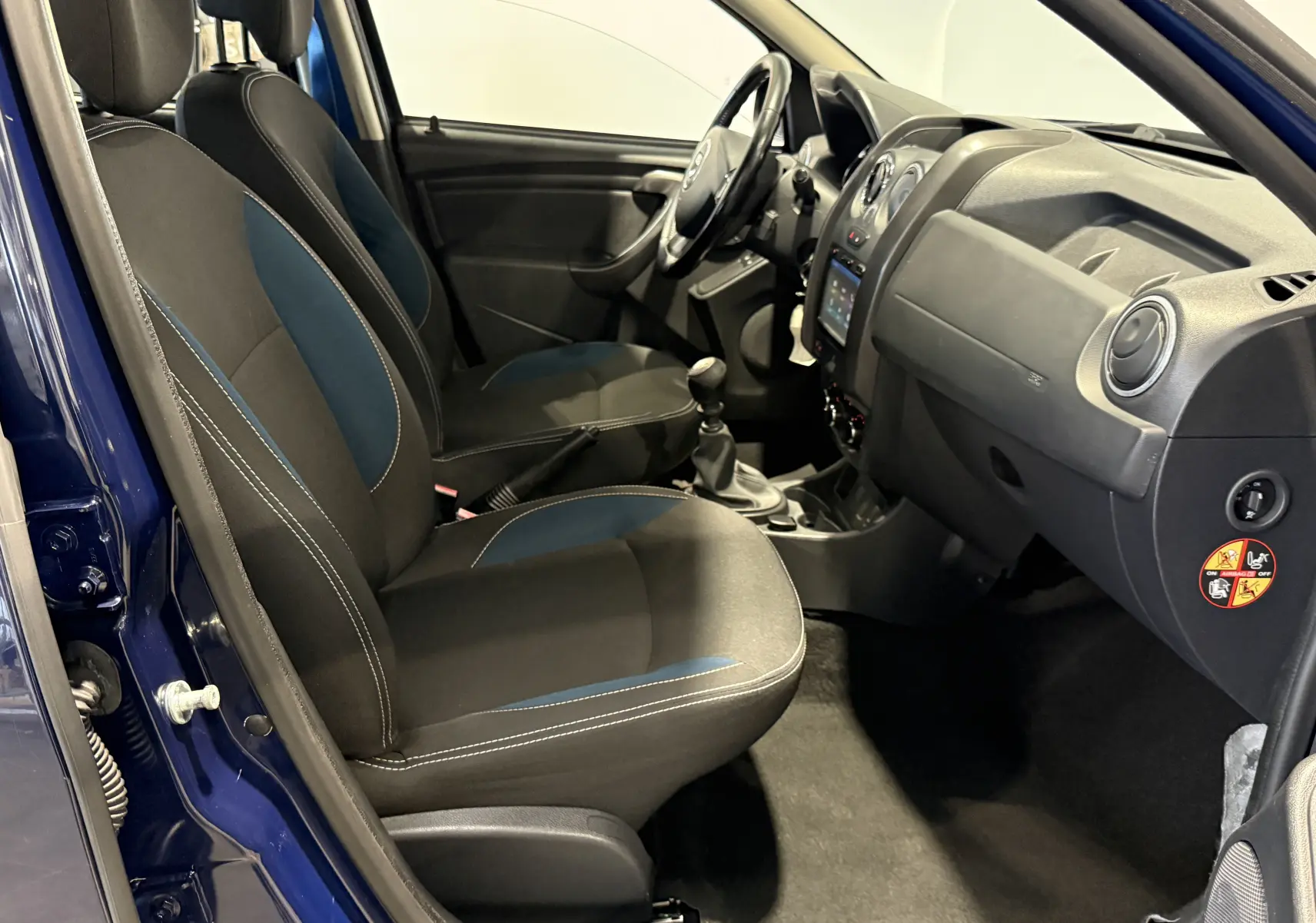 Vue intérieure côté conducteur du Dacia Duster bleu 2015, montrant sièges avant, tableau de bord et levier de vitesse manuel.