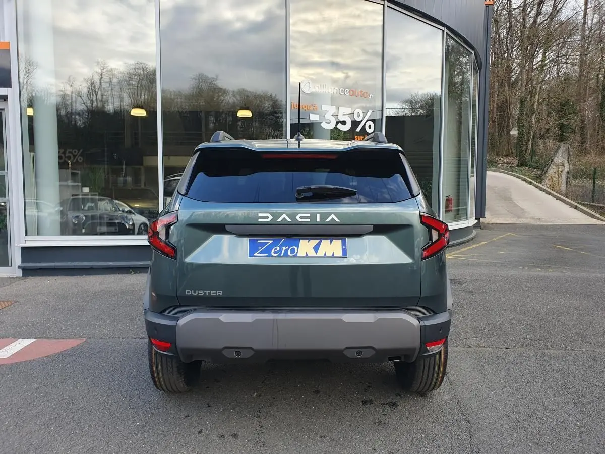 Vue arrière d'un Dacia Duster 2026 vert cèdre avec barres de toit et feux LED distinctifs, stationné devant une vitrine.