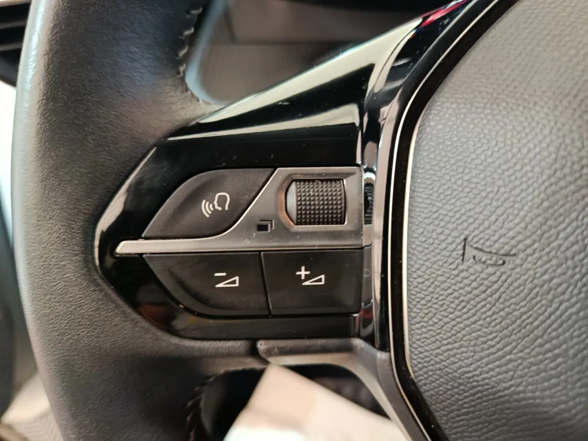 Gros plan sur les commandes gauche du volant cuir noir de la Peugeot 208 blanc, avec boutons de volume et reconnaissance vocale.