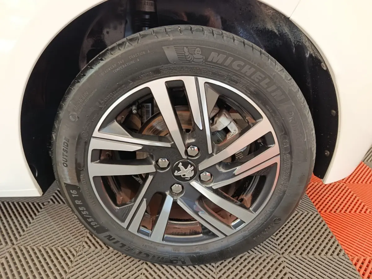 Gros plan sur la roue avant droite d'une Peugeot 208 blanche, mettant en valeur la jante alliage noire et argent.