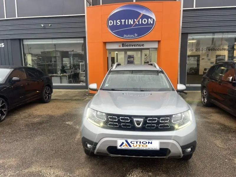 Vue frontale d'un Dacia Duster 1.5 Blue dCi gris Comète métallisé avec barres de toit, phares allumés, devant un garage.