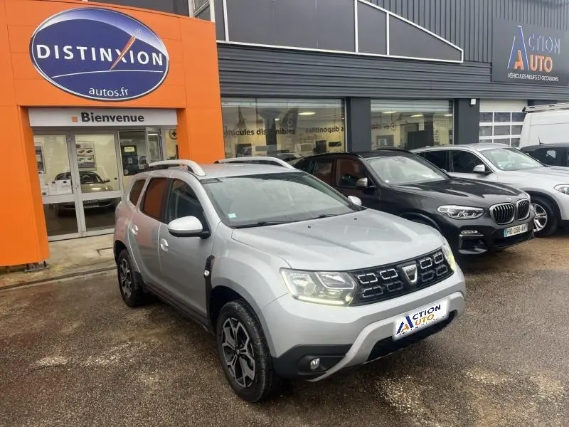 Vue 3/4 avant d'un Dacia Duster gris Comète métallisé garé devant un concessionnaire avec barres de toit et jantes alu noires.